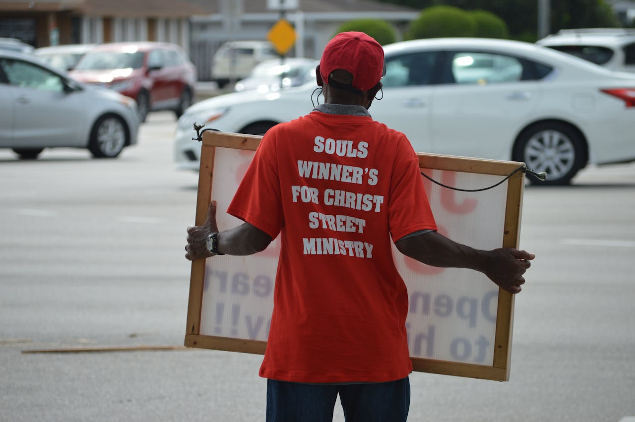 A man in red shirt with Christian message holding a sign on a busy street in Florida.