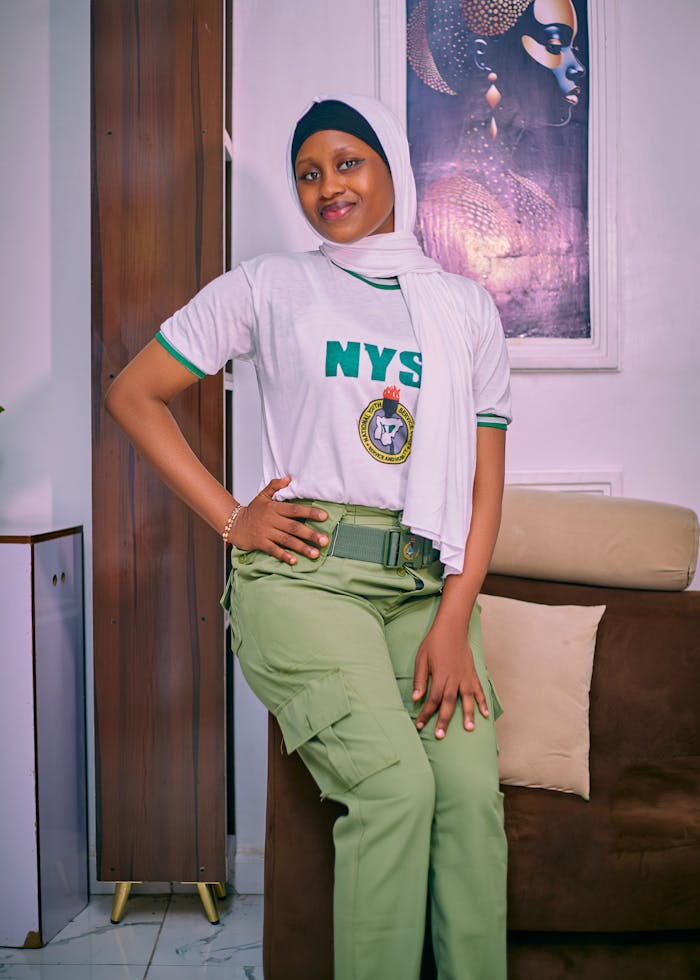Smiling young woman in NYSC uniform posing confidently indoors. Captured in a lively, warm setting.